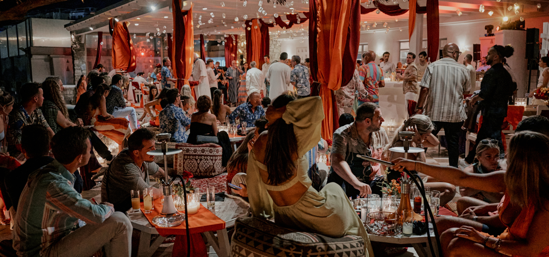 Wedding guests enjoying the vibes of a henna pre wedding party, decorated venue with warm colors and textiles on this arabian party in Mykonos