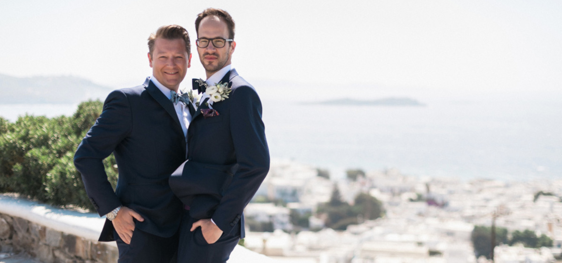 Two men in suits on a ledge, enjoying the view of the sea during a Mykonos same-sex wedding celebration.
