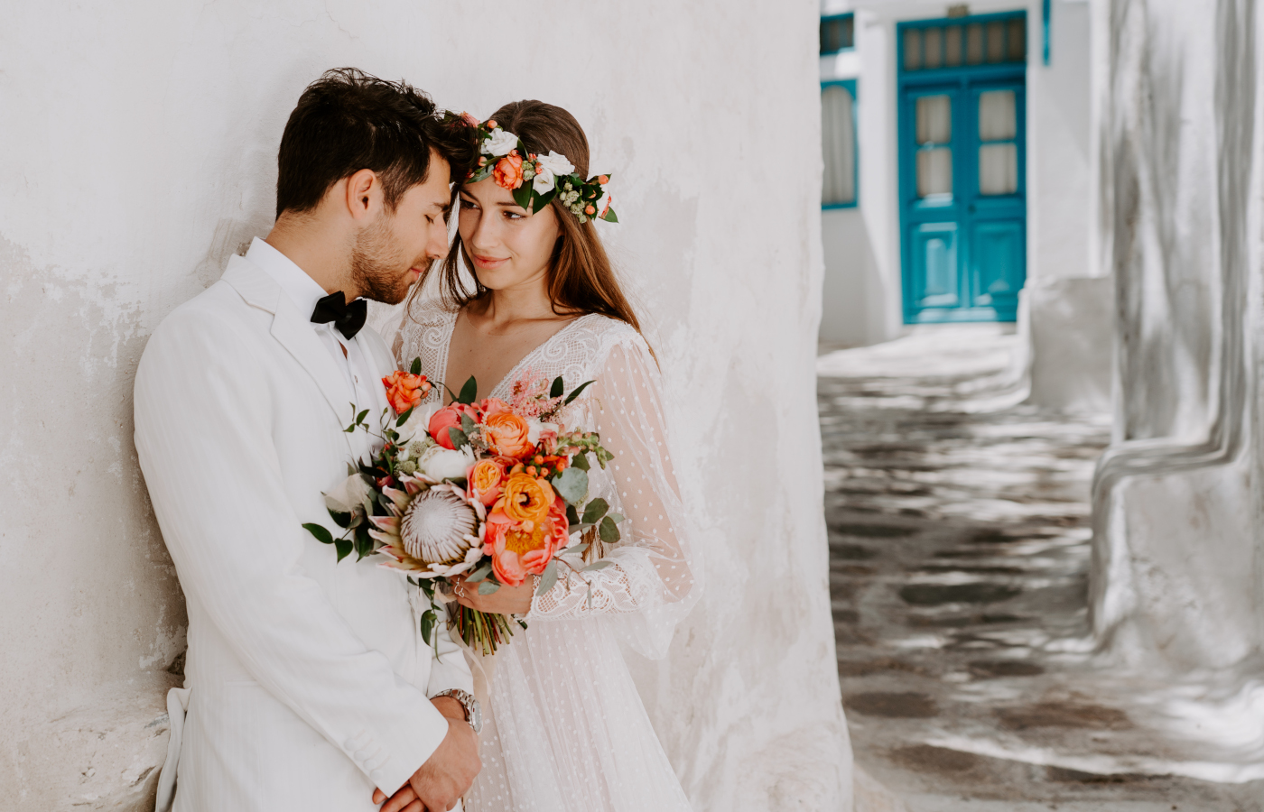 Bride and groom leaning on a white wall in the streets of Mykonos Town, bride is holding a coral bridal bouquet and wearing a coral flower head crown.